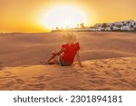 A tourist woman with a sunhat in Maspalomas Dunes of Gran Canaria sunset. She either listens to distant ocean waves or watches sand dunes shift in the breeze while closing her eyes.