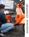 Small photo of Yogyakarta, Indonesia - Dec 31 2022: A man sits beside his bright orange vespa strada aom motorcycle while smoking. Translate: mbayung green lombok stall benny, skilled in jogja