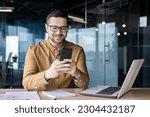 Small photo of Smiling young male office worker sits at a desk with a laptop and writes a message on the phone, chats, checks mail, reads the news, break at work.