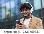 Close-up photo. Young smiling Indian male student standing outside wearing white headphones and holding phone. Calls, chats, listens to music.