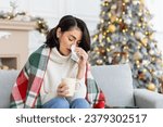 Small photo of A young woman fell ill during the Christmas and New Year holidays. She sits at home on the sofa covered with a blanket, wipes her nose from a runny nose with a napkin, holds a cup of drink, medicine.