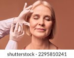 Small photo of Aesthetic medicine.Portrait of an adult woman with closed eyes on a beige background. Hands in white gloves and a medical gown make an injection in the cheekbone.