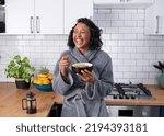 A young multi-ethnic woman laughs while eating breakfast cereal in dressing gown