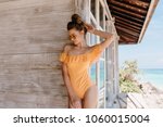 Happy lightly-tanned woman touching her dark hair while standing near wooden wall. Outdoor shot of enchanting brunette girl in sunglasses and orange swimwear.