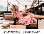 Ecstatic blonde girl playing with her hair while chilling in cafe. Inspired young woman in striped dress enjoying champagne in restaurant in summer weekend.