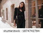 Beautiful young brunette with bright makeup, dark dress, black jacket, belt and bag posing on street near beige building and window in sunny city