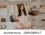 Thoughtful brunette woman in pink pajamas looks at sandals. Lady with red lips poses in dressing room and choose between two pairs of high heels.