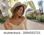 Sunny image of young stylish woman standing on street, in fashionable hat close-up. She has gentle smile and closed eyes. Nice neckline and bare shoulders.