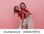 Small photo of Beautiful young asian girl hugs her caucasian girlfriend after long absence indoors. Blonde glasses in red T-shirt and white pants along with brunette denim dress. Summer playful mood concept