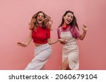 Small photo of Wonderful young racial girls are dancing to beat of music on pink background in studio. Pretty women in colorful casual clothes are smiling broadly holding hands. Merry holiday concept
