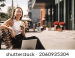 Small photo of Asian brunette woman with wide smile sitting on bench looks at camera with coffee in her hands on city street. Girl manager in non-office is engaged in additional work at laptop.