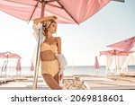 Beautiful caucasian woman is standing on beach crowded with sun loungers with umbrellas. Gorgeous blonde with bandana on her head in yellow swimsuit and white shirt looks to side.
