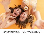 Small photo of Close-up of cheerful young caucasian female friends fooling around, posing for camera, on yellow background. Smiling broadly, long-haired brown-haired women lie relaxed on floor.