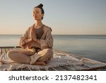 Relaxed young caucasian woman sitting on seashore practices yoga without stress. Model with dark topknot on her head with eyes closed. Cozy beach atmosphere, summer concept.