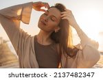 Close-up of young caucasian relaxed woman touching her hair in bright sunlight. Brown-haired girl with closed eyes, wearing brown T-shirt and blouse on top. Good mood, fashion trends