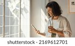 Small photo of Positive young brunette caucasian woman smiles looking at her smartphone screen indoors. Hottie wears casual clothes while sipping juice. Mood, lifestyle, concept .