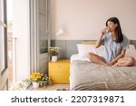 Nice young caucasian girl drinks water, crossing legs sitting on bed in room. Woman with brunette hair closes her eyes in pleasure. Good morning concept