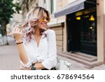 Pretty girl wearing sunglasses and bracelets playing with her short curly hair and smiling on the street. Outdoor portrait of laughing blonde young woman in white shirt standing near store.