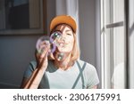 Young woman in cap blowing soap bubbles next to the window. Concept: fun, house, objects