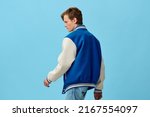 a handsome, young man student in a trendy blue bomber jacket stands with his back to the camera on a light blue background.