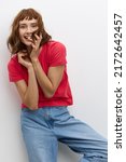 Small photo of vertical photo of a funny, mischievous woman in a red T-shirt standing on a white background and covering her face with her hair, leaning on the wall