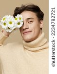 Small photo of Close-up photo.A beauteous young man with shining skin, with dark, short, beautiful hair combed back, in a beige turtleneck with a high collar stands on a dark beige background with a bouquet of