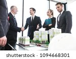 Small photo of Business people standing near residential quarter houses models and shaking hands, meeting of architects and investors