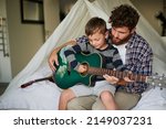 Small photo of String by string. Cropped shot of an adorable little boy sitting on his dads lap while learning to play the guitar at home.