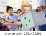 Small photo of Teacher, recycle bin and kid in classroom throwing trash for cleaning, climate change or eco friendly in school. Recycling plastic, sustainable learning or education with boy student in kindergarten.