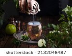Small photo of Woman hand pouring Green Apple Irish Mule cocktail cocktail from shaker in copper mug surrounded by ingredients and bar tools on dark wooden table surface