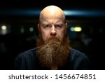 Small photo of Bearded man with shaved head staring at the camera with an ill-tempered frown and cross expression lit from above at night in a close up portrait