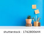Home office background. Desk with notes, supplies, design objects, plant, wood elements and blue wall.