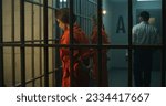 Small photo of Two female prisoners, inmates in orange uniforms stand facing the metal bars in front of prison cells. Prison officer walks, watches women criminals in jail. Detention center or correctional facility.