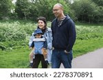 Small photo of A smiley multicultural family walking on a footpath in Figgate Park in spring season. The mother is carrying her infant son with a baby carrier. The baby wears a misplaced hat and carries a toy.