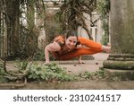 Small photo of Irish female new yoga teacher doing an eight-angle pose wearing orange sportswear in the terrace of a landscaped garden in Hampstead Heath, London. Plants and kiwis on the floor. Looking at camera.