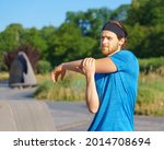Young handsome man athlete warming up, stretching arm before morning workout in green park, sportive male in blue shirt and headband exercising outdoors on sunny day. Sport and people concept