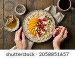 Faceless female holding ceramic plate with oat porridge with sliced peach, raspberries, chopped almond, coconut chips, chia seeds, honey and tea. Traditional breakfast served on wooden background