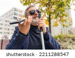 Elderly gray-haired caucasian entrepreneur with low vision in sunglasses sitting on bench, having a phone call using speaker mode, and holding white walking crane with one hand. Outdoor closeup shot