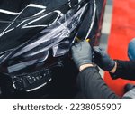Small photo of Two hands in protective black rubber gloves covering vehicle headlight with film to darken them. Closeup indoor shot. Car detailing. High quality photo
