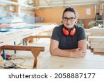 Small photo of Happy confident woman carpenter or woodworker in a factory leaning on a workbench with ear muffs around her neck smiling at the camera