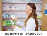 Small photo of Single pharmacy technician in white lab jacket holding prescription scrip and generic medication box with shelf in background