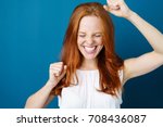 Attractive young redhead woman celebrating a victory punching the air with her fists and a beaming toothy smile over a blue studio background with copy space