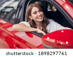 Cheerful young female sitting in shiny red car on passenger seat and looking out open window while enjoying purchase in dealership