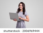 Happy adult business lady in elegant dress looking at camera while working on laptop against gray background