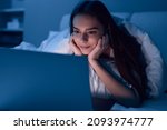 Young ethnic female with long dark hair lying on bed with hands on cheeks while watching interesting movie on laptop at night at home