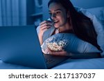 Happy young woman eating popcorn while lying on bed under blanket and watching movie on netbook at night at home