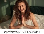 Small photo of Smiling female with closed eyes and clasped hands praying while feeling gratitude and sitting on bed in morning at home