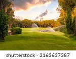 Small photo of Watering grass in golf course at sunset with beautiful sky. Scenic panoramic view of golf fairway. Golf field with pines