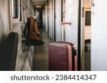Small photo of Red suitcase in the train. Baggage standing in the corridor of night sleeping train, Nobody. Empty wagon, cozy and comfortable travel, sunset light coming from the window.