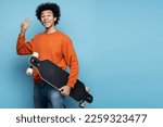 Handsome smiling African American man holding longboard showing victory sign isolated on blue background. Portrait of attractive happy skater looking at camera, copy space 